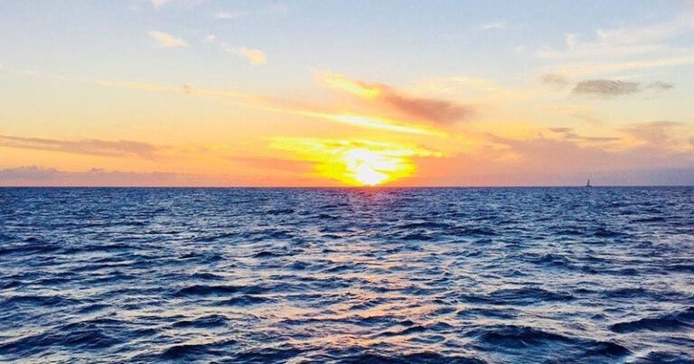 The sun setting on the open ocean with a Waikiki Sunset Cruise in the distance.